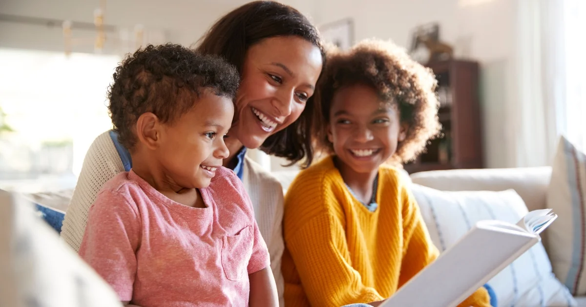 Smiling mother reading a book with her two happy children on a couch at home.